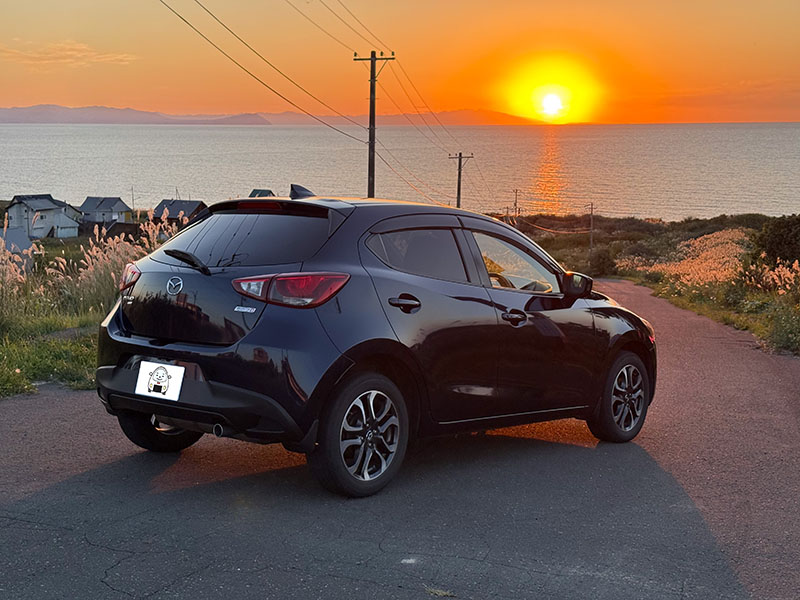 夕日の海辺に停めたマツダ デミオ XDツーリング 4WD（後ろ斜めから）